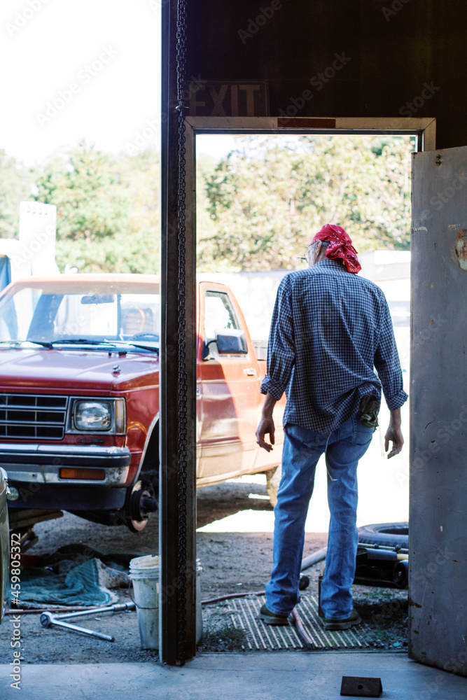 An older man mechanic standing in his auto shop garage doorway looking out at red pick up truck and salvage junk yard