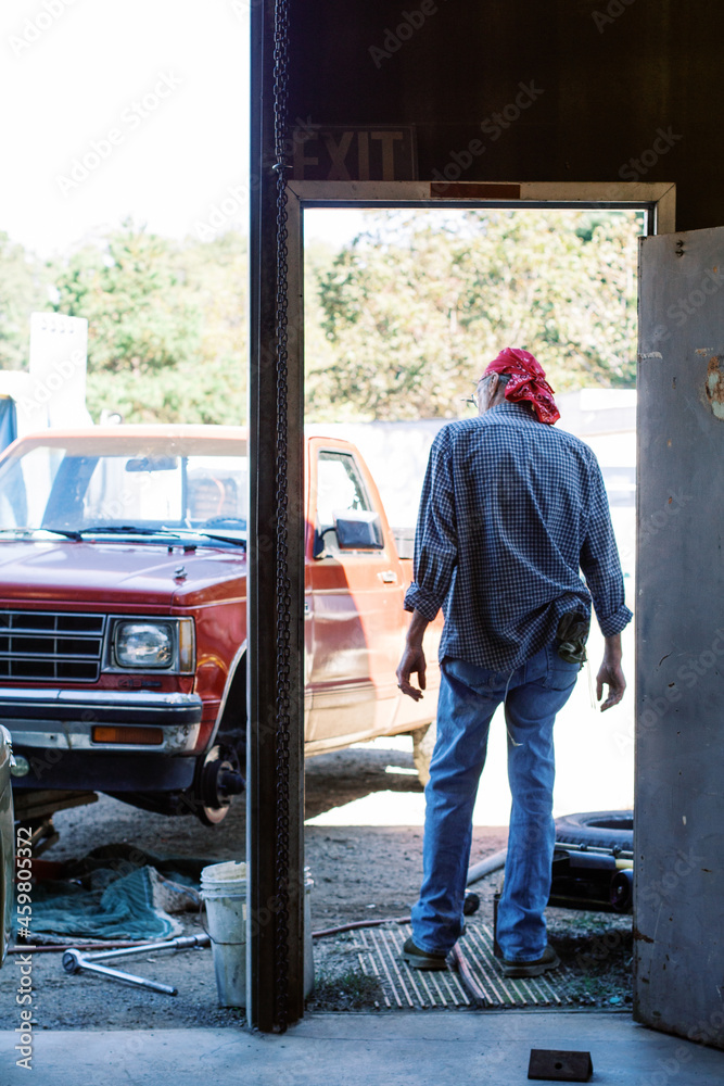 An older man mechanic standing in his auto shop garage doorway looking out at red pick up truck and salvage junk yard An older man mechanic standing in his auto shop garage doorway looking out at red pick up truck and salvage junk yard