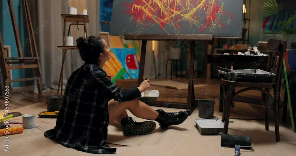 A woman is working in her studio, school, and art class on a new piece of painting related to abstractionism, modernity. The girl crouches on the floor picks up a tray and thinks about the color A woman is working in her studio, school, and art class on a new piece of painting related to abstractionism, modernity. The girl crouches on the floor picks up a tray and thinks about the color