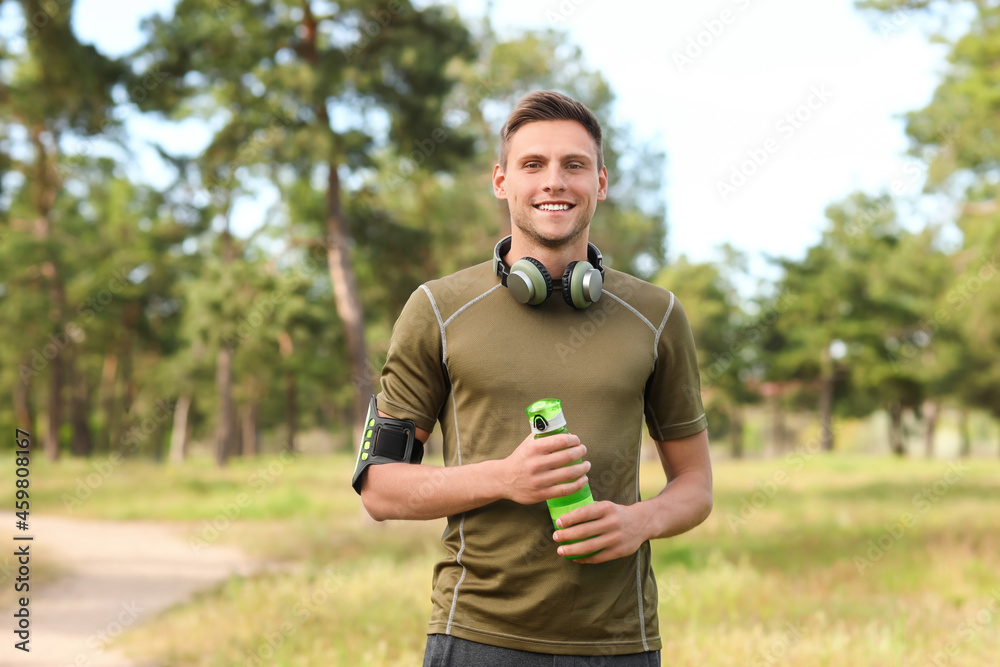 Sporty young man with bottle of water and headphones in park - obrazy, fototapety, plakaty 