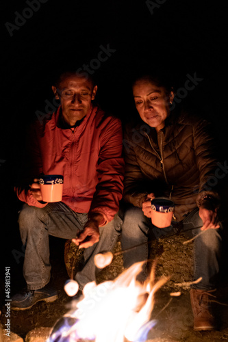 A couple camping, holding cups in front of a campfire