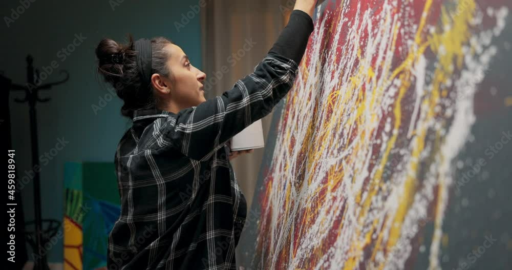 A student wearing a plaid shirt during an art class is painting an abstract, modern painting with blue paint that she holds together with a brush in her hands.