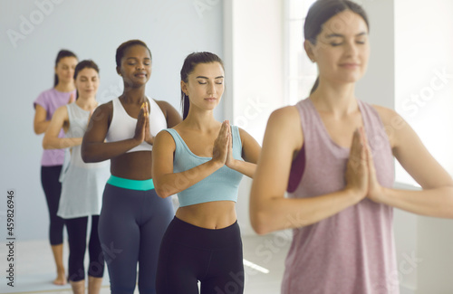 Diverse group of happy relaxed young women having class at yoga studio. Team of beautiful slender ladies in fitness clothes standing in gym, doing Namaste with eyes closed, enjoying breathing exercise