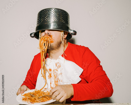 Man making a mess eating pasta with sauce while using a colander as a hat