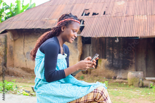 A happy woman or lady sitting and using a smart phone as she sits outside a village mud house