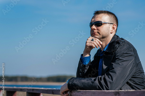 A young man in a black coat and sunglasses stands on an old wooden bridge and looks into the distance. A young, handsome businessman on the riverbank. 