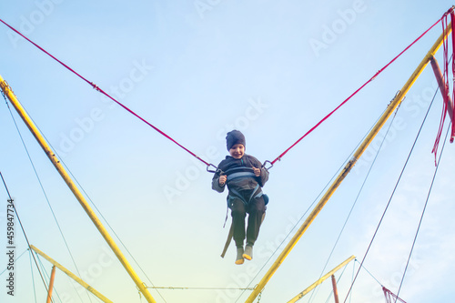 The boy is jumping on a bungee trampoline. A child with insurance and stretchable rubber bands hangs against the sky. The concept of happy childhood and games in the amusement park. © Siniehina