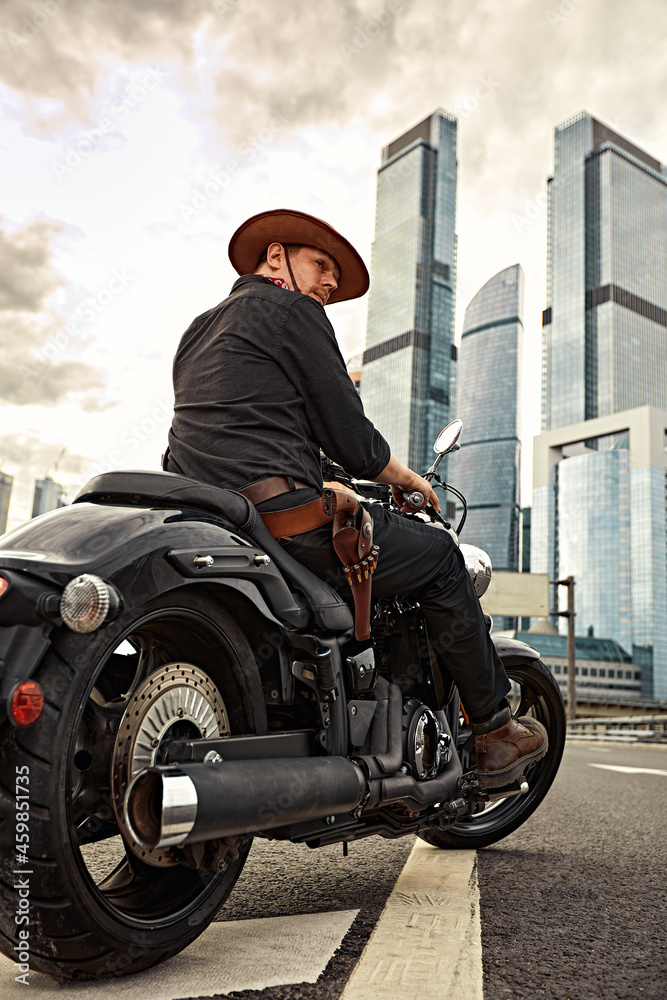 Fototapeta premium Portrait of stylish male dressed in a black leather jacket and bandana with gun sitting on his custom-made retro motorcycle, looking away, buildings of the big city on background.