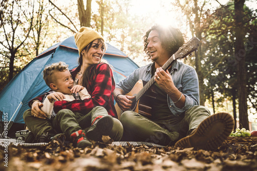 Happy family camping in the forest playing guitar and singing together - Mother, father and son having fun trekking in the nature sitting in front of the tent - Family, nature and trekking concept