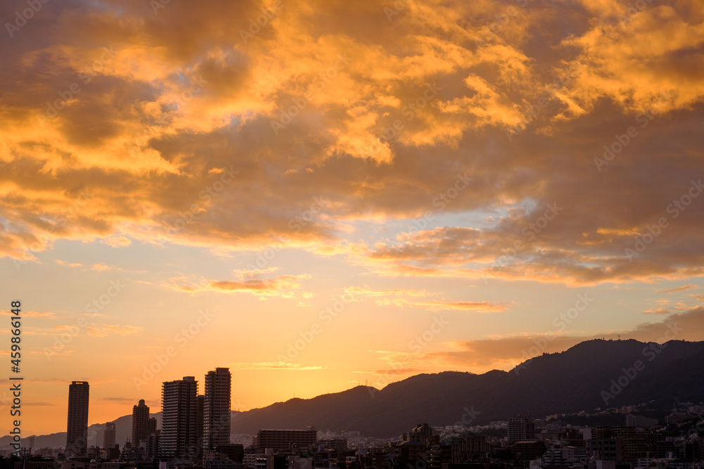 Fototapeta premium 都会の夕暮れ。六甲山に沈む夕陽とオレンジ色に染まる空と雲。高層ビルがシルエットで浮かぶ。