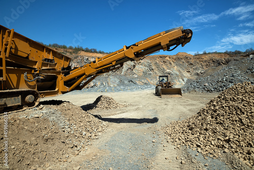 A stone crusher machine and a bulldozer in an open-type stone quarry on a sunny day.