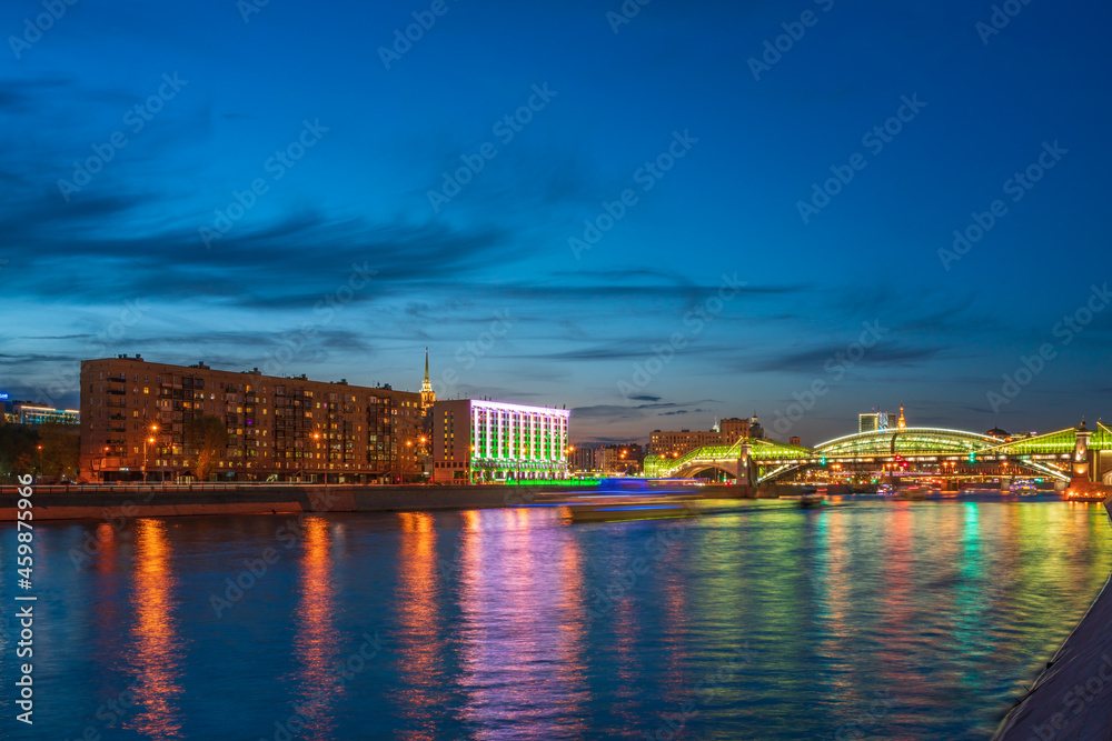 View of the colorful Bogdan Khmelnitsky bridge illuminated at night reflecting in the Moskova river. Moscow Kiyevsky railway station at night. Moscow, Russia
