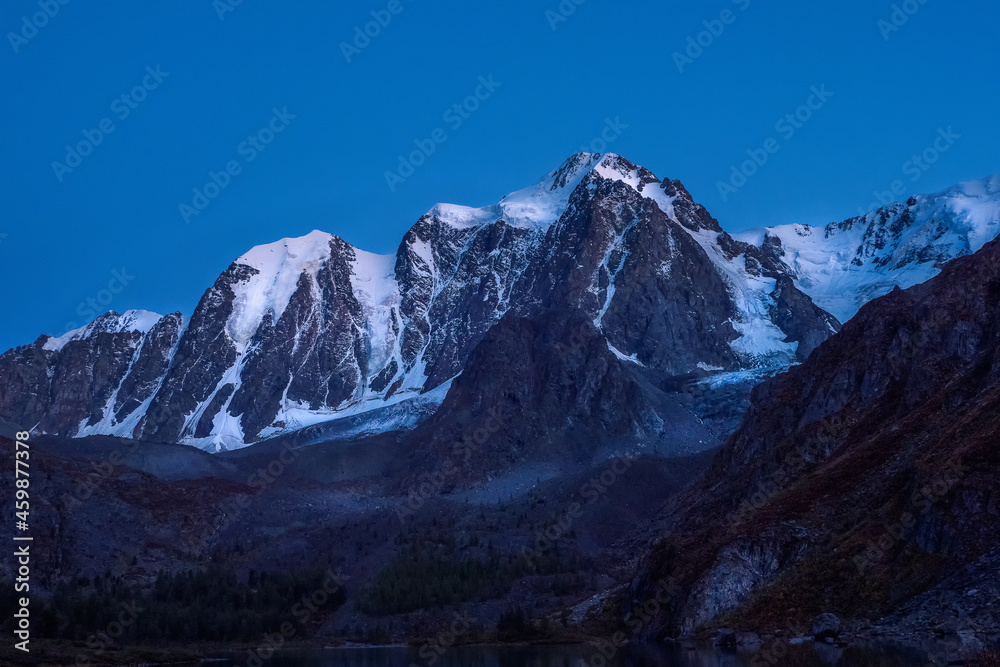 High snowy mountains illuminated by the light of the moon in a clear sky at dusk.