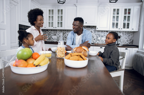 African american parents and their two little daughters sitting on modern kitchen  eating and drinking. Positive family of four enjoying tasty breakfast together at home.