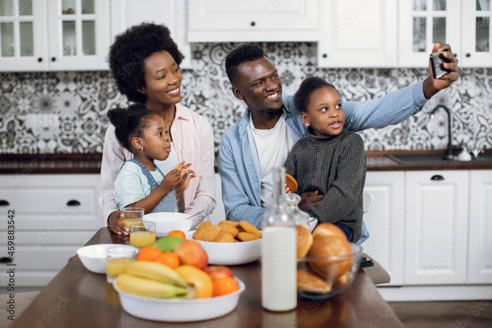 Beautiful african family taking selfie while sitting on kitchen with fresh fruits and cookies. Young parents with two pretty daughters using modern smartphone during breakfast at home.