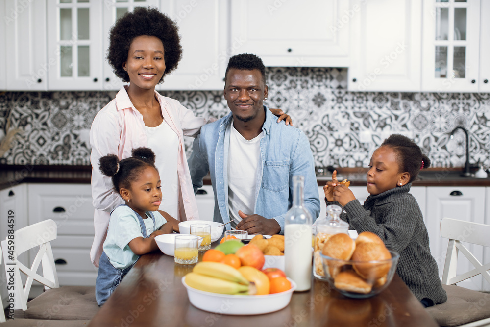Front view of young african parents with two pretty daughters sitting at kitchen table with fresh fruits, juice and cookies. Breakfast time of happy family. Domestic lifestyles. Front view of young african parents with two pretty daughters sitting at kitchen table with fresh fruits, juice and cookies. Breakfast time of happy family. Domestic lifestyles.