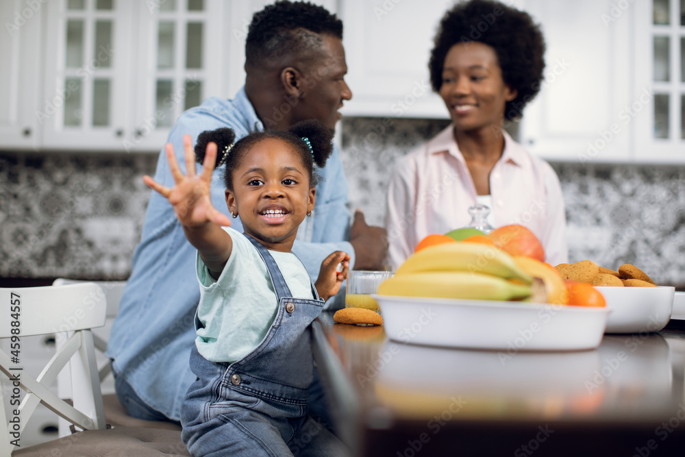 Pretty smiling girl waving hand on camera while sitting at modern kitchen with her parents. African wife and husband talking on blur background. Happy family spending time together at home. Pretty smiling girl waving hand on camera while sitting at modern kitchen with her parents. African wife and husband talking on blur background. Happy family spending time together at home.