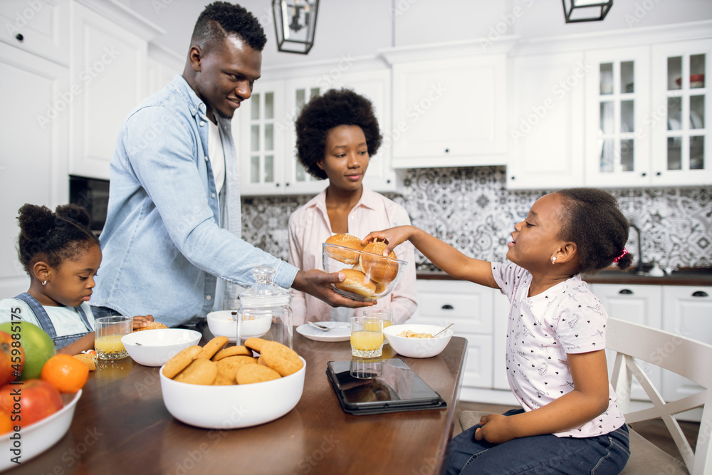 Young african parents and their cute daughter having breakfast with fruits and freshly baked buns. Cute little daughter takes tasty bun from the bowl, looking at her father