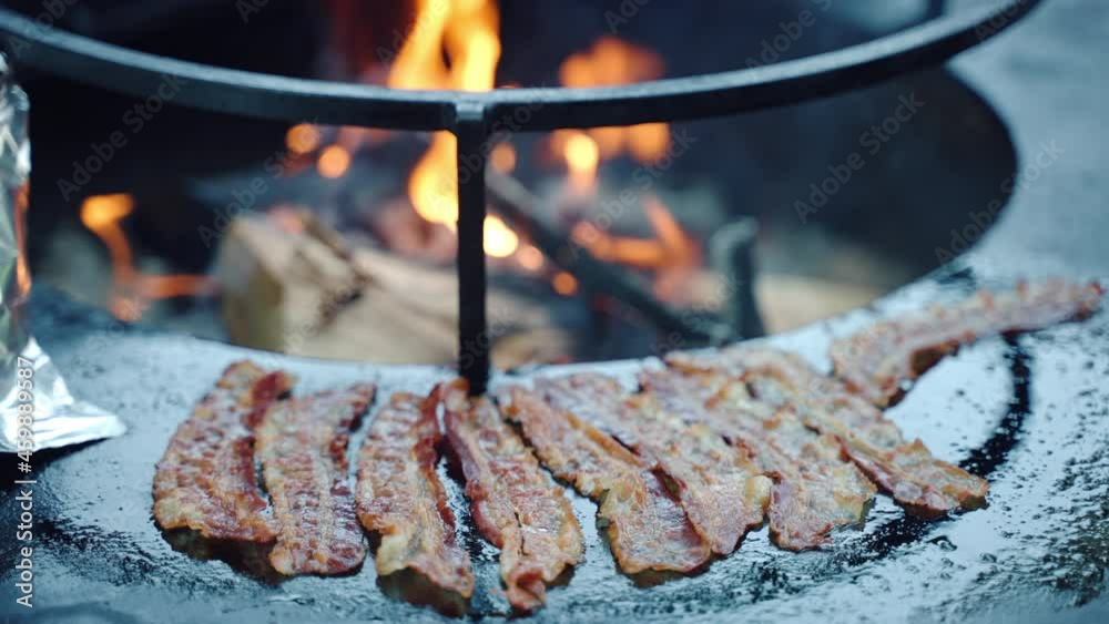 Traditional barbecue picnic party outdoor, thin crispy bacon slices grilling on bbq smoker grid with burning wood fire. Process of preparation weekend picnic with wide range of meat, unhealthy eating