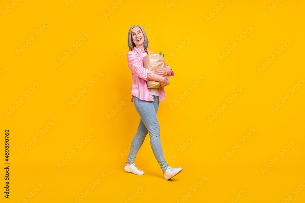 Full length profile side photo of aged woman happy positive smile go hold shopping bag food isolated over yellow color background Full length profile side photo of aged woman happy positive smile go hold shopping bag food isolated over yellow color background