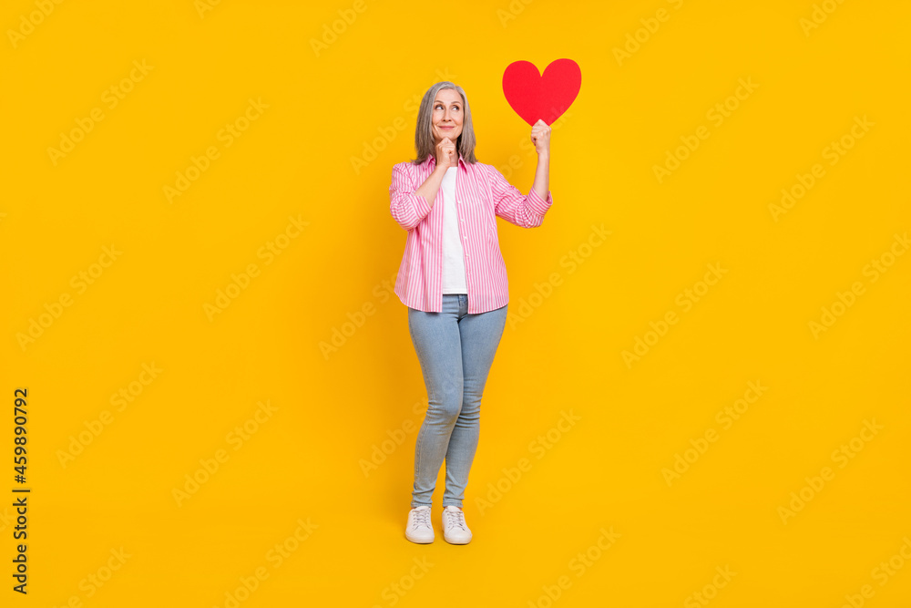 Full body photo of mature woman happy positive smile think hold paper heart love isolated over yellow color background Full body photo of mature woman happy positive smile think hold paper heart love isolated over yellow color background