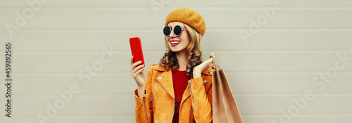 Autumn portrait of happy smiling young woman taking a selfie by smartphone with shopping bags wearing an yellow jacket, beret on gray background