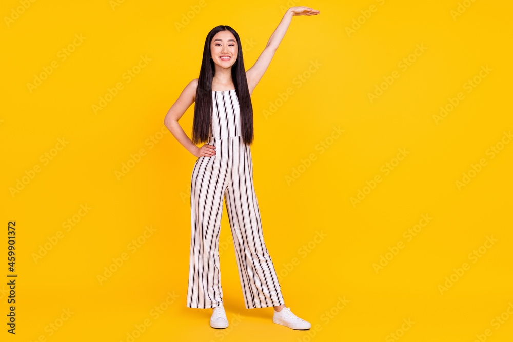 Full length body size photo girl smiling in striped overall showing height with hand isolated vibrant yellow color background