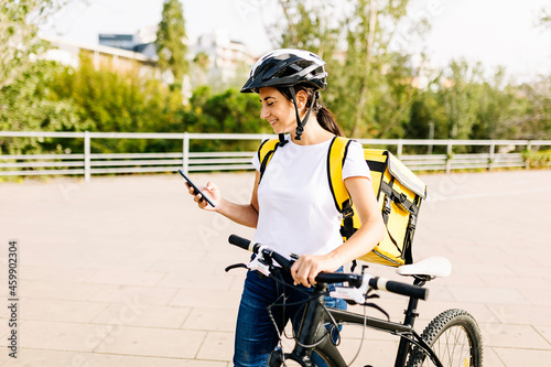 Smiling delivery woman using mobile phone while wheeling bicycle