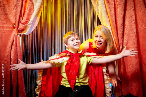 Family during a stylized theatrical circus photo shoot in a beautiful red location. Models mother and son posing on stage with circus curtain