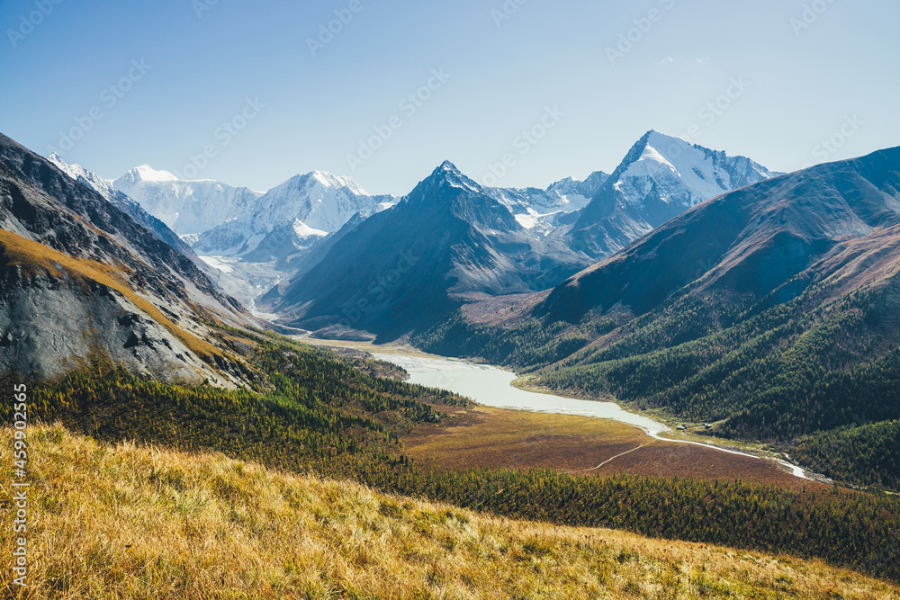 Fototapeta premium Wonderful alpine landscape with mountain lake and mountain river in valley with forest in autumn colors on background of snowy mountains silhouettes under blue sky. Beautiful mountain valley in autumn
