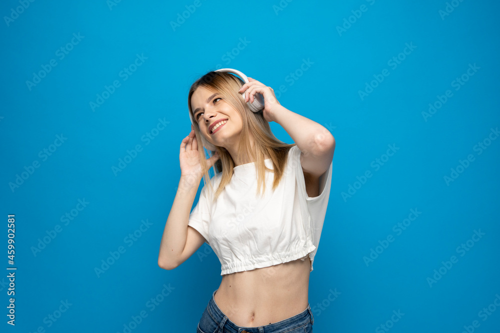 Beautiful attractive young blond woman wearing white t-shirt and glasses in white headphones listening music and dancing on blue background in studio. Relaxing and enjoying. Lifestyle.