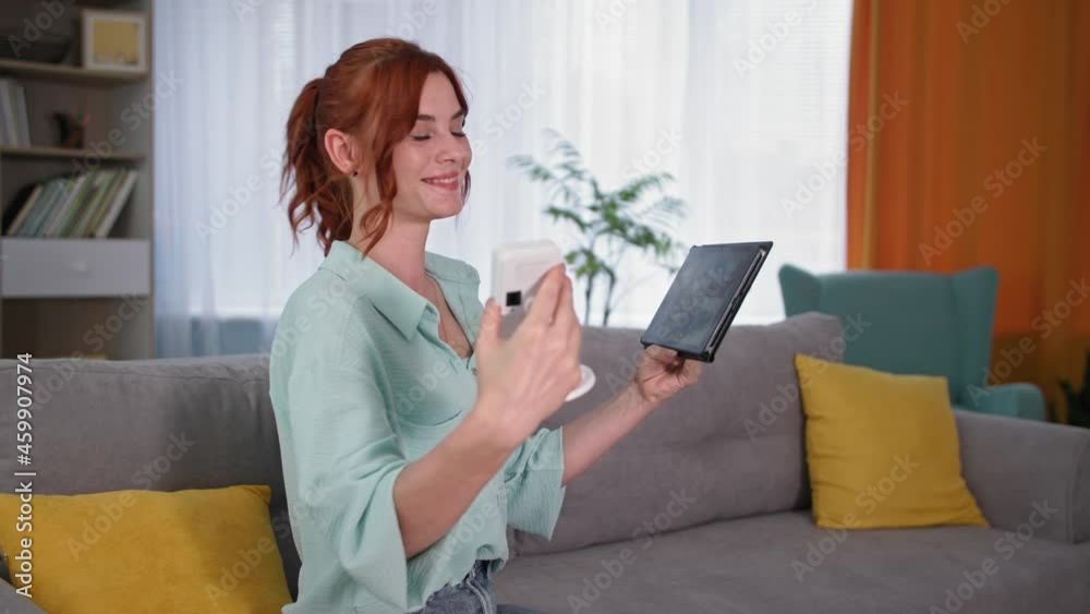 home security app, smiling female houseowner sitting on the couch with surveillance camera and tablet in hands home security app, smiling female houseowner sitting on the couch with surveillance camera and tablet in hands