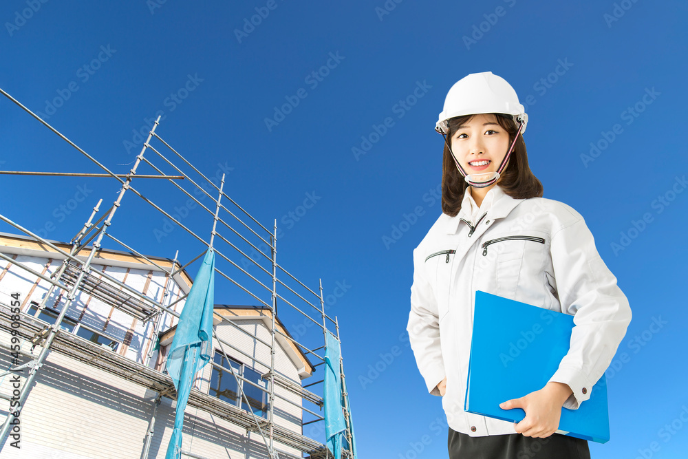 青空と一戸建て工事現場を背景に立つ、作業着を着たカメラ目線の女性