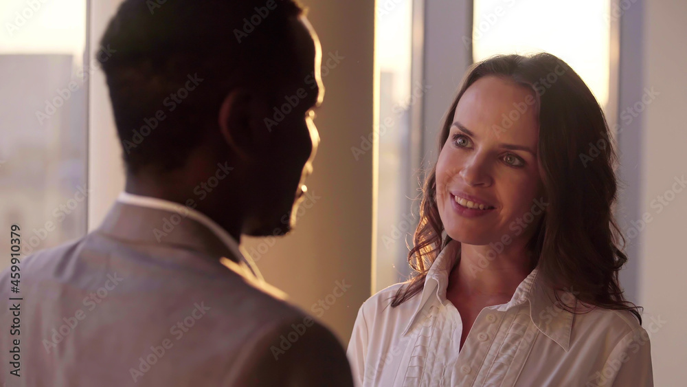 Shot of two smiling mixed colleagues standing by large panoramic window and talking Shot of two smiling mixed colleagues standing by large panoramic window and talking