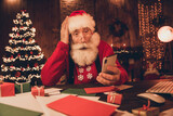 Photo of aged santa claus amazed shock hand touch head look read browse cellphone noel new year magic lights indoors