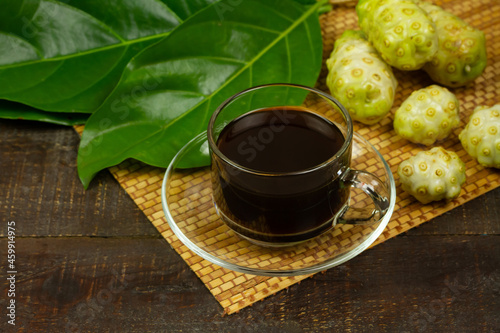 Noni juice in transparent glass and noni fruit with green leaf on wooden background. 