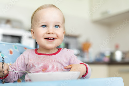 Cute happy adorable infant baby boy enjoy having fun sit in chair learning eat with spoon and plate healthy vegetable soup puree at home kitchen. Children feeding healthy food nutrition diet concept
