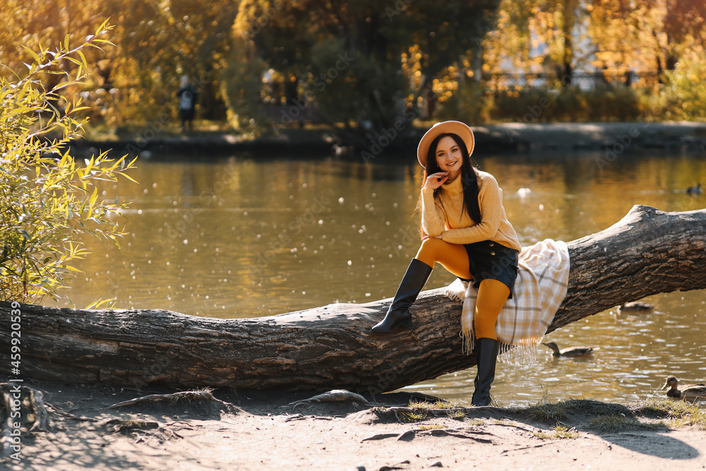 A pretty young woman in a yellow sweater and a fashionable hat is sitting relaxing on a tree near the water in the autumn forest outdoors, selective focus