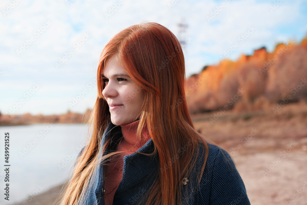 portrait of happy young red haired woman smiling and looking aside. local travel. spending time on a beach, relaxing and having fun. woman wearing blue coat.