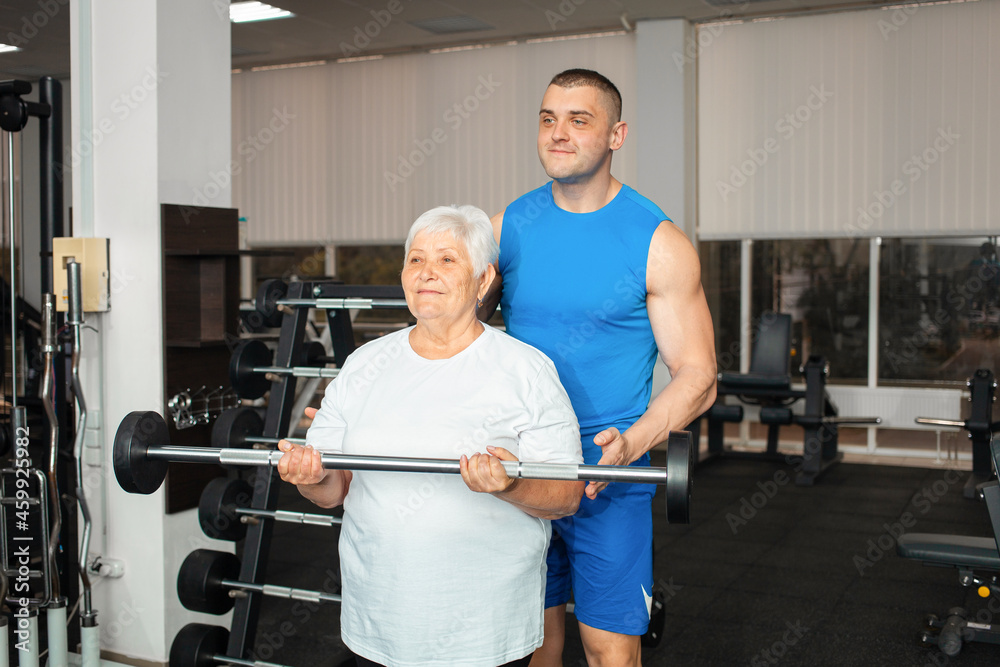 pensioner, an elderly woman performs an exercise with a barbell in gym. coach helps and shows. Healthy sports lifestyle, senior concept. woman with gray hair is recovering from injury. Happy pensioner, an elderly woman performs an exercise with a barbell in gym. coach helps and shows. Healthy sports lifestyle, senior concept. woman with gray hair is recovering from injury. Happy