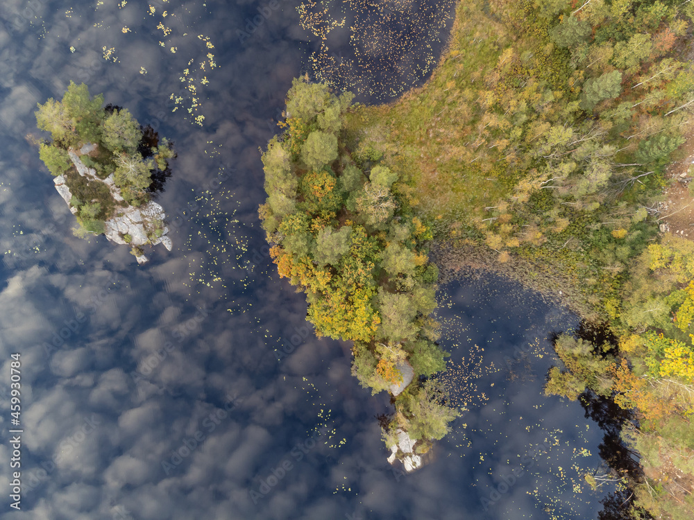 Drone nature photography taken from above in Sweden in fall time. Bird's eye view of a lake with trees in autumn colors and a small island. Dark blue water surface with reflections from clouds.