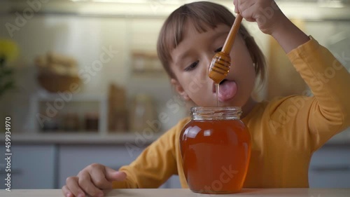 ittle child girl eat honey as it drops on her tongue licking it out from the wooden spoon. Kid eats honey from a jar at home in the kitchen. Healthy organic sweets for kids