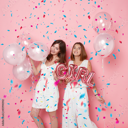 Portrait of happy young women, dressed in a white, holdings a balloons, over colored confetti, pink background.