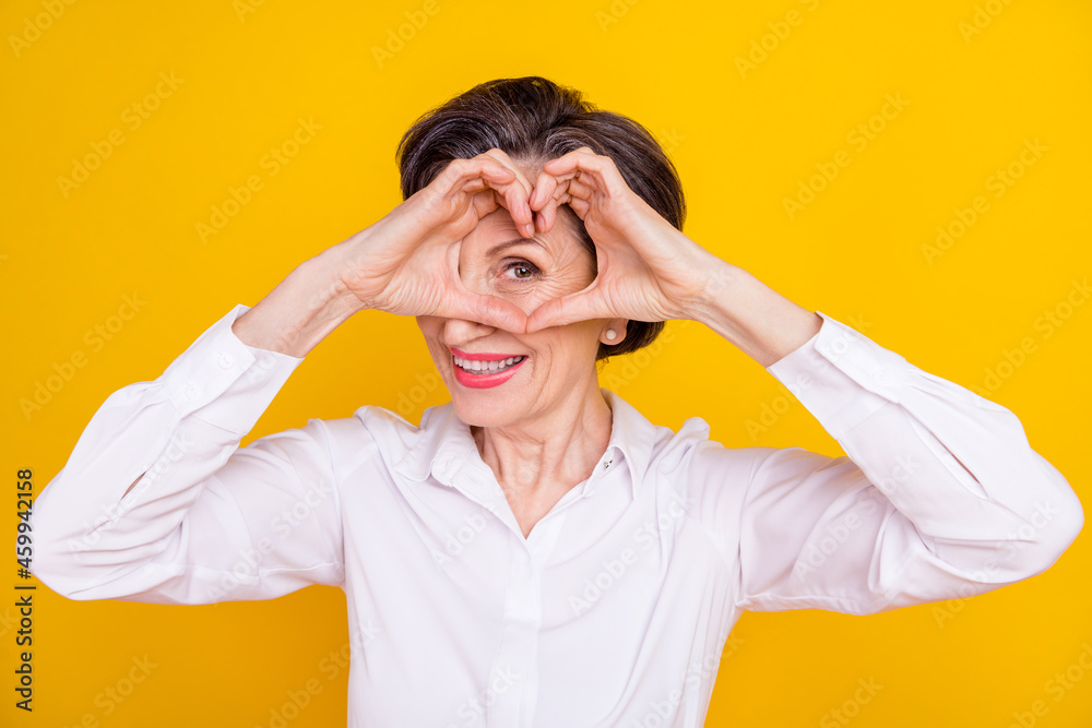 Photo of cute adorable age woman dressed white shirt showing fingers heart cover eye smiling isolated yellow color background Photo of cute adorable age woman dressed white shirt showing fingers heart cover eye smiling isolated yellow color background