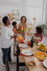 Smiling african american woman holding turkey near family and thanksgiving dinner