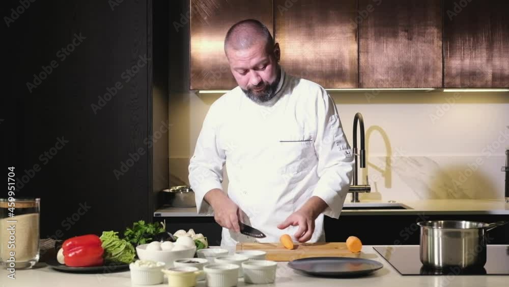 Man in an apron in the kitchen is preparing a homemade lunch. The cook cuts fresh juicy carrots with a sharp knife for a delicious dish. Preparation of ingredients Man in an apron in the kitchen is preparing a homemade lunch. The cook cuts fresh juicy carrots with a sharp knife for a delicious dish. Preparation of ingredients