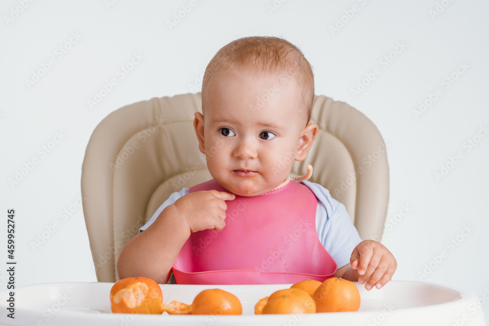 A baby in a pink bib sits on a chair and points a finger questioningly at himself. Lots of tangerines on the table in front of him A baby in a pink bib sits on a chair and points a finger questioningly at himself. Lots of tangerines on the table in front of him