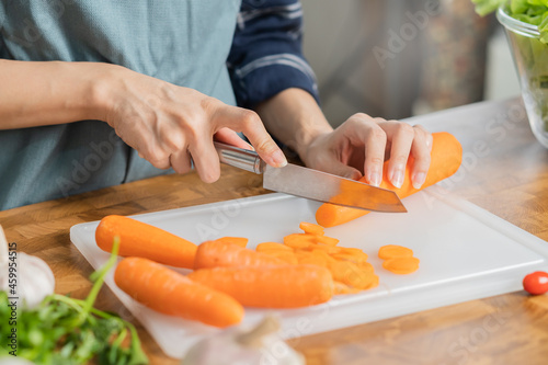 Asian young woman, girl or housewife using knife, cutting carrots on board, on wooden table in kitchen home, preparing ingredient, recipe fresh vegetables for cooking meal. Healthy food people.