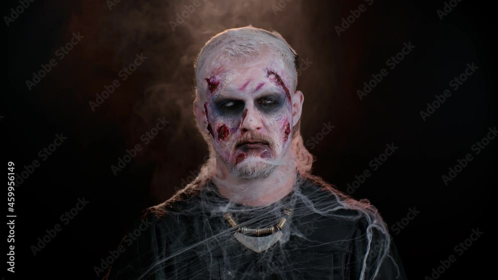 Sinister man with horrible scary Halloween zombie makeup in costume making faces, looking ominous at camera, trying to scare. Dead guy with wounded bloody scars face isolated against black background