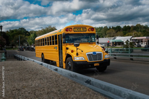 A school bus in Upstate NY, Chenango Broom County, crosses a bridge in town on its way to pick up Children at the end of a school day.  Yellow bus, white clouds, blue sky in Autumn.